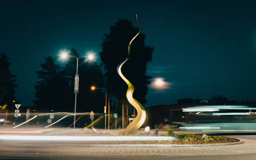 Kuvassa Ihana-taideteos illalla ja ohi ajavia autoja sekä linja-auto